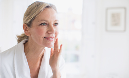 Smiling mature woman applying moisturizer to face at bathroom mirror