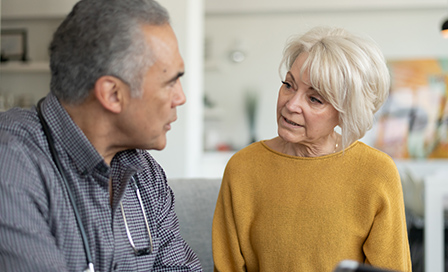 A doctor and an elderly patient are indoors at the woman's home. The doctor is talking to the woman while holding a tablet computer. She looks slightly concerned.