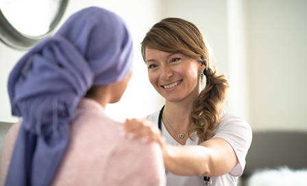 A young woman is indoors in a hospital. She has cancer, and her head is covered with a scarf. She is sitting next to her female doctor. Her female doctor is trying to discuss outcomes with her patient while comforting her.