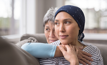 An ethnic woman wearing a headscarf and fighting cancer sits on the couch with her mother. She is in the foreground and her mom is behind her, with her arm wrapped around in an embrace, and they're both looking out the window in a quiet moment of contemplation.