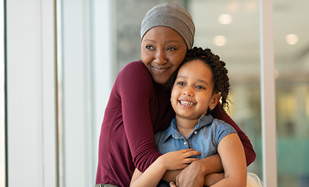 A beautiful woman with cancer takes a break from chemotherapy treatment at the hospital. She is seated near a large bay of windows with her young daughter. The two are embracing and looking out the window. They are smiling and filled with hope, gratitude and happiness