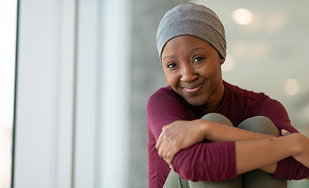 A beautiful woman with cancer wears a hat. She is seated near a large bay of windows and her arms are wrapped around her knees. She is taking a break from receiving chemotherapy treatment. The woman is smiling at the camera. She appears to be at peace and hopeful.