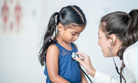 A young hispanic girl looks down at the stethoscope that her doctor is placing on her chest. Her doctor is a young caucasian woman and is wearing professional clothing.