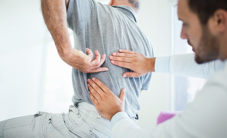 Closeup rear low angle view of an early 60's senior gentleman having some back pain. He's at doctor's office having medical examination by a male doctor. The patient is pointing to his lumbar region.