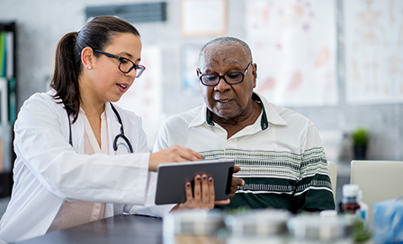 A senior man of African descent is indoors in a hospital room. He is watching his female doctor using a tablet computer. She is explaining a medication schedule to him.