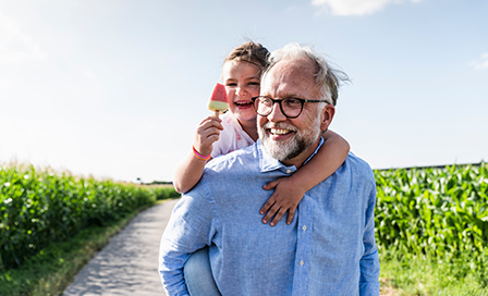 Father carrying his daughter on his back, walking down a dirt road surrounded by cornstalks.